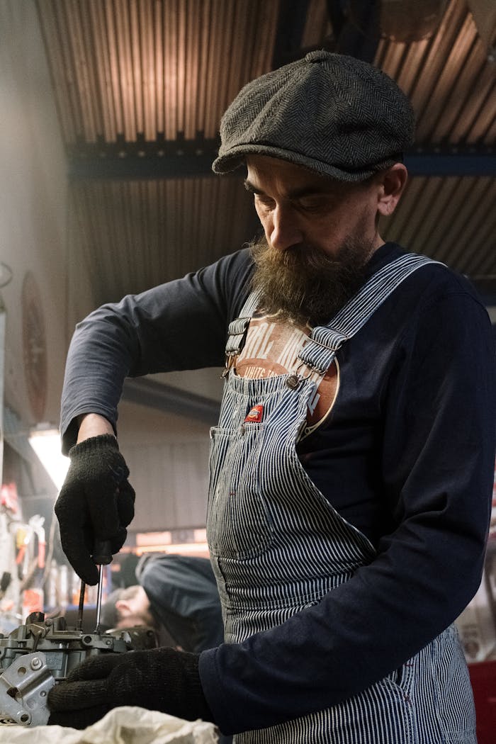 Bearded mechanic in striped overalls repairing a vehicle engine inside a workshop.