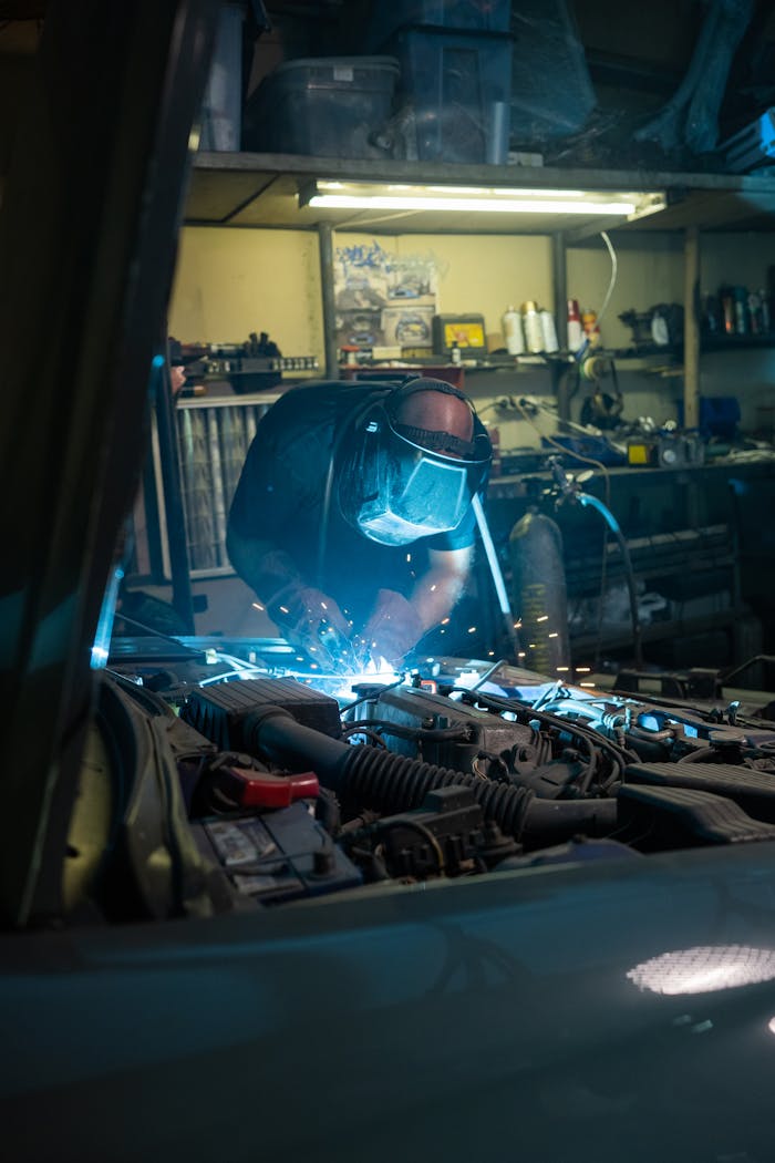Mechanic wearing safety gear welding under hood of a car in a garage workshop.